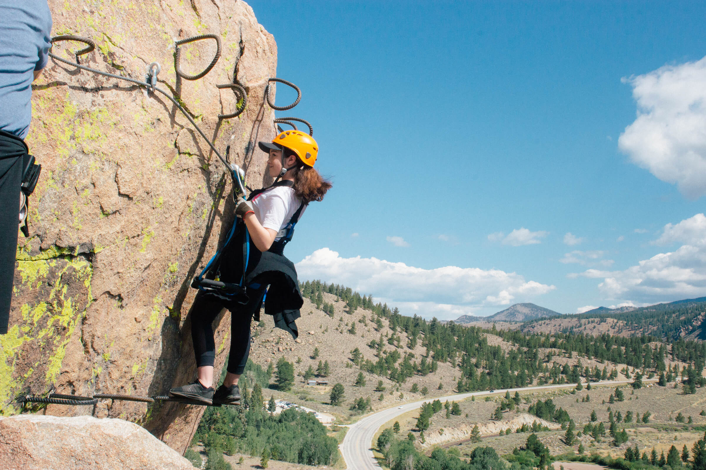 A woman in an orange helmet traversing across a rock face with a Colorado view in the background